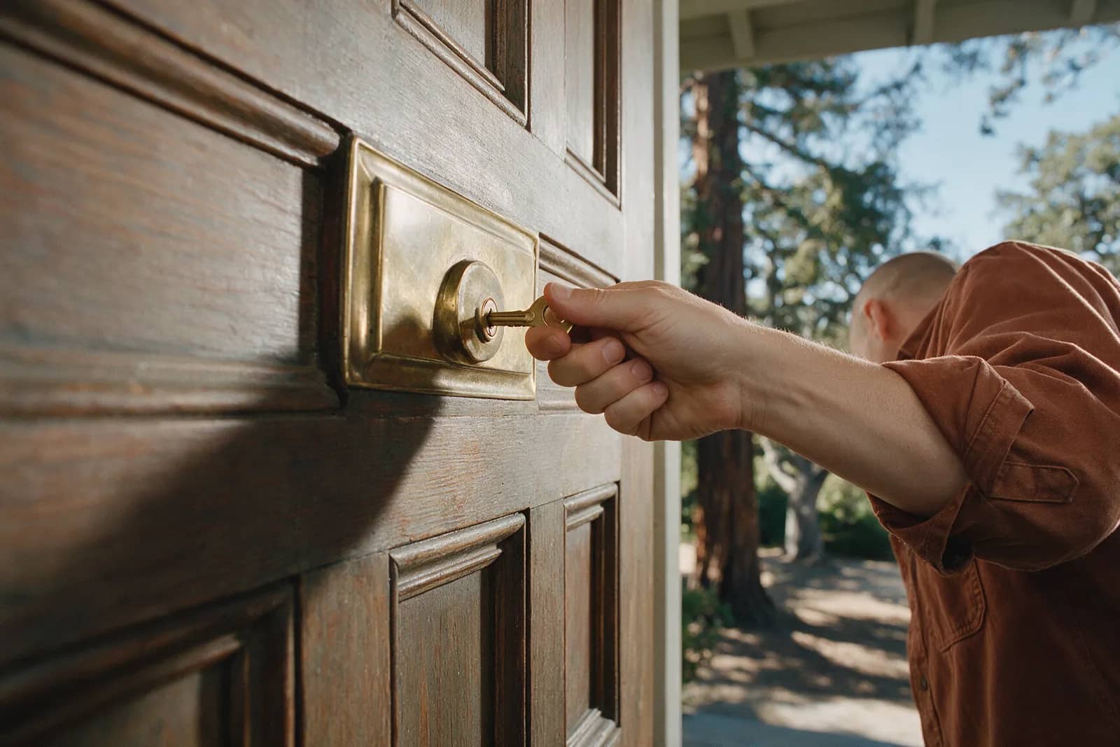 A hand turning a brass key in the brass escutcheon of a dark paneled front door, redwoods on the path behind